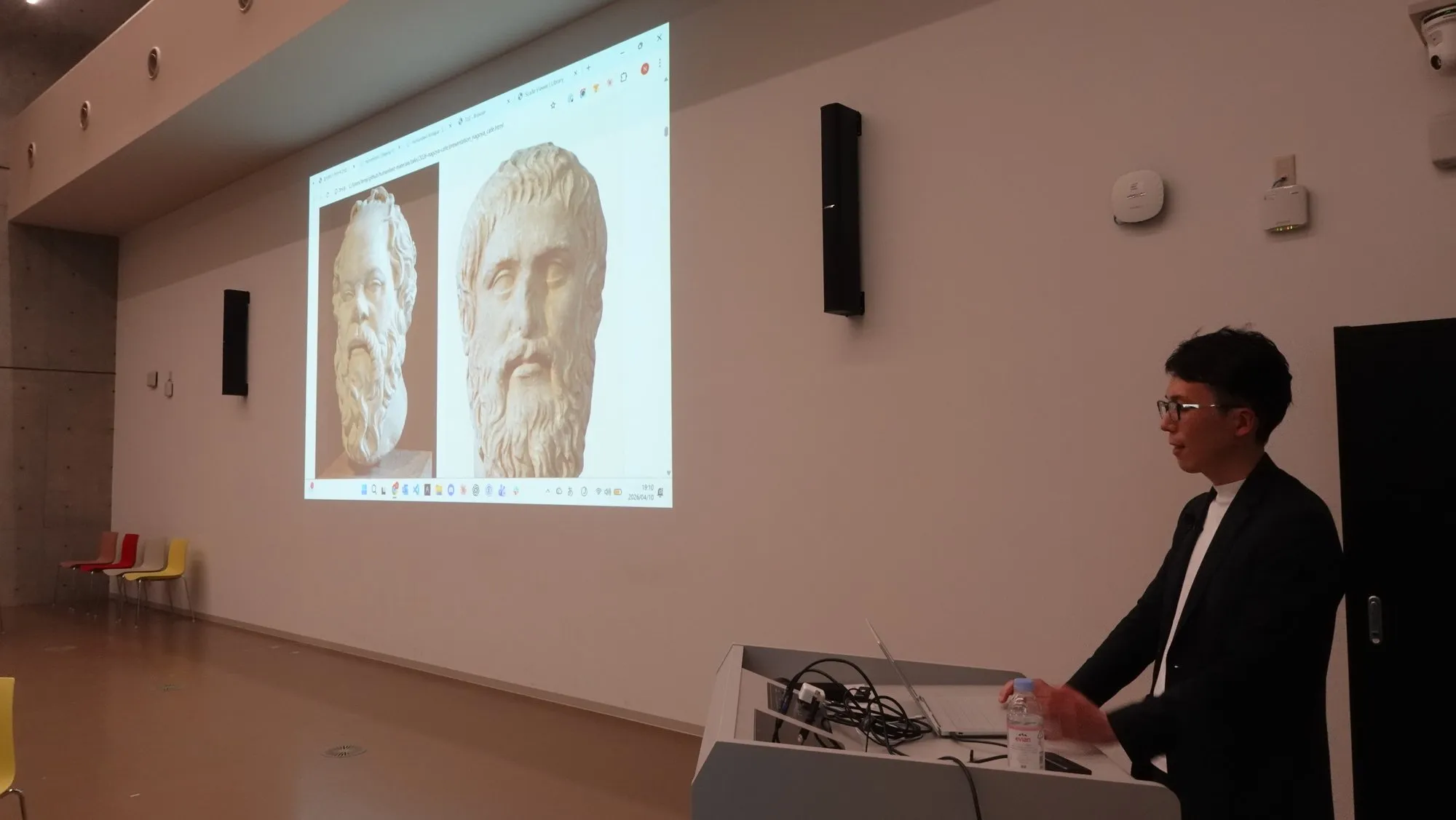 Naoya Iwata speaking at the 127th NU Cafe, with busts of Socrates and Plato on the screen